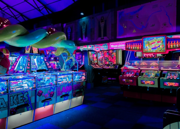 An indoor arcade filled with vibrant gaming machines lit with neon blue and pink lights. The scene includes various arcade games with flashing screens and colorful displays, and a large overhead structure resembling a green snake. The background shows illuminated signs and themed decor, contributing to an immersive entertainment atmosphere.