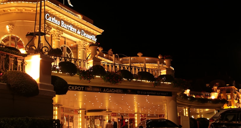 A luxurious casino at night, illuminated with warm, golden lights. The building features elegant architecture with columns and decorative elements. A few people are walking towards the entrance, and there are a couple of parked cars nearby.