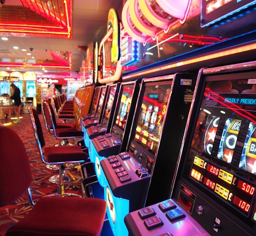 A brightly lit casino interior featuring a row of colorful slot machines. The neon lights create a vibrant atmosphere, with patterned carpeting adding to the decor. Stools are aligned neatly in front of each machine.