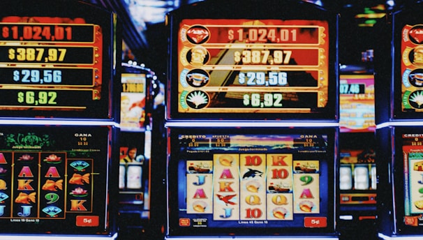 A row of colorful slot machines lit with bright neon lights, showcasing various graphical elements on the screens, indicating different games and jackpots. The atmosphere suggests a lively casino setting, bustling with excitement.
